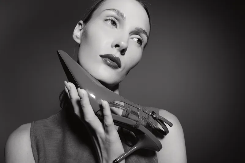 Portrait of a pale skinned Caucasian woman holding a high heel shoe near her face. Concept of fashion, elegance, and beauty in black and white photography.
