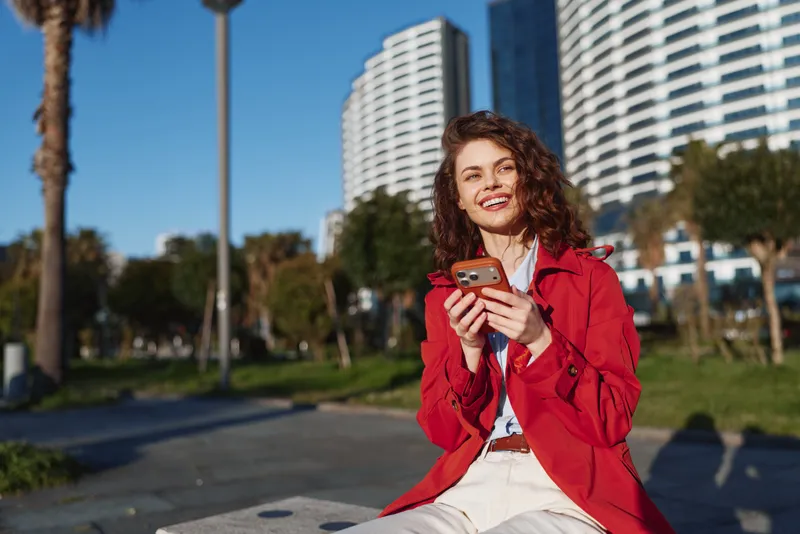 A young woman with curly hair sits on a park bench in an urban plaza, smiling while checking her smartphone.