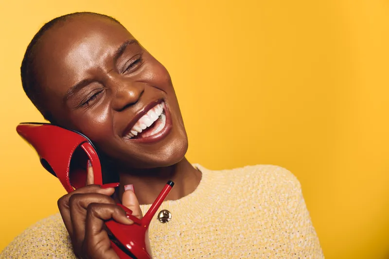 Happy African woman with dark skin smiling and holding red high heel shoe near face on yellow background. Concept of joy, fashion, beauty, and playful mood in studio.
