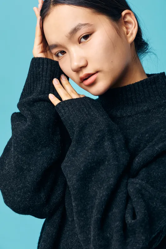 Young woman posing with hand near face wearing black oversized sweater standing against blue background with serious expression and natural makeup for portrait.