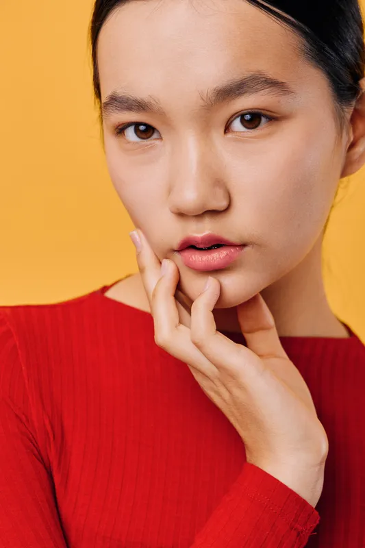 Young woman with clear skin and dark hair wearing red top posing with hand near face on yellow background. Closeup, natural makeup and thoughtful expression with studio lighting.
