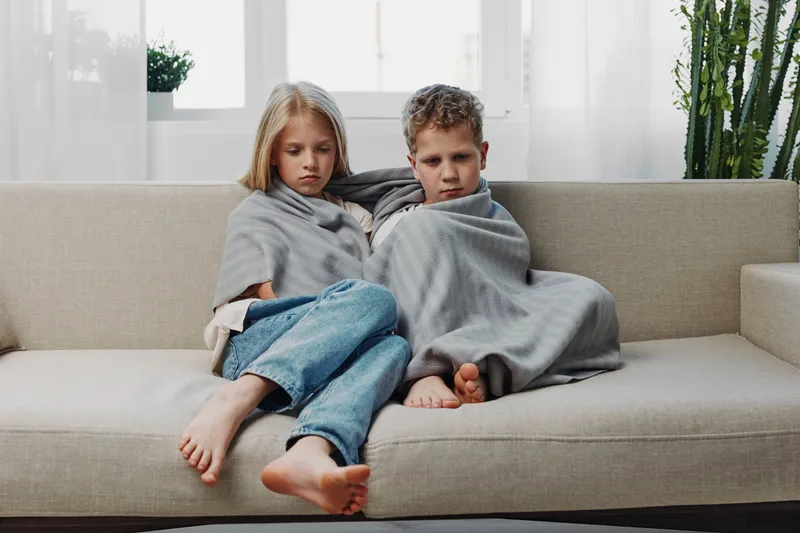 Sad brother and sister wrapped in gray blankets sitting barefoot on beige sofa in bright room. Concept of cold, illness, family care, and comfort during recovery or difficult times.