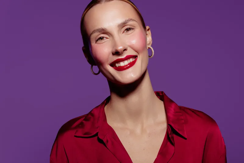 Smiling woman with red lipstick and gold earrings wears red blouse against purple background. Studio portrait with bright makeup and confident expression. Concept of beauty and fashion.