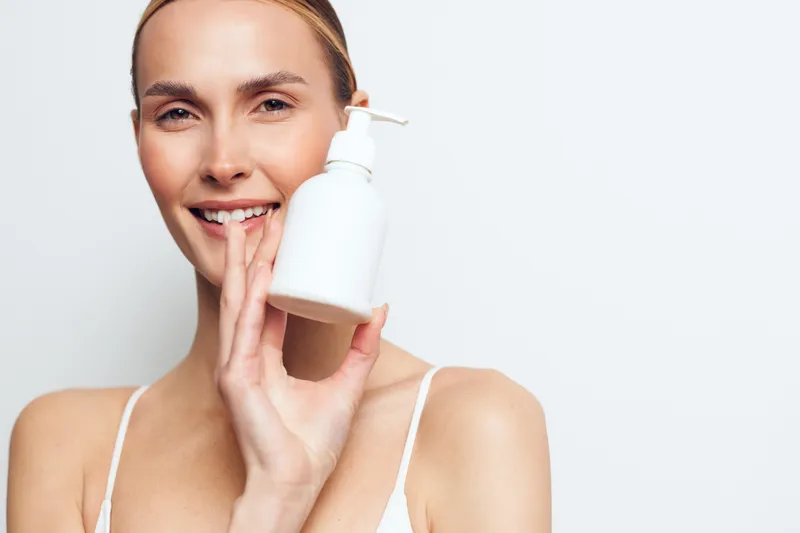Smiling woman with clear skin holding white pump bottle near face on white background. Concept of skincare, beauty, self-care, and wellness with soft natural lighting and fresh mood.