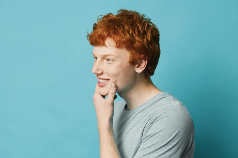 Young man with red hair smiling and looking sideways, hand on chin, wearing gray t shirt against blue background in studio portrait with copy space for text.