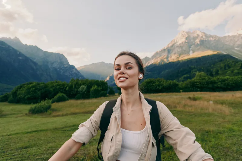 Smiling woman with backpack in a mountain meadow, embracing nature, outdoor lifestyle and travel. Bright natural light highlights wellness and happiness in the scene.