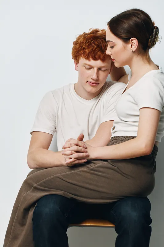 Young couple with woman sitting on man's lap, holding hands and showing affection in casual white t shirts and neutral background in studio portrait.