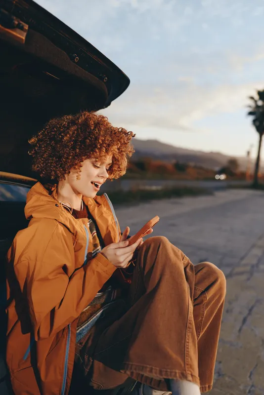Outdoor woman lifestyle scene, curly haired woman smiles near open car trunk, orange jacket and casual clothing, hand gesture while standing on street sidewalk at sunset