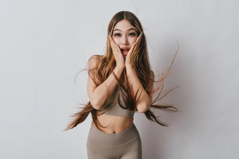 Young woman with long hair and surprised facial expression standing against white background. She wears beige fitness clothing and holds her hands on cheeks with wide eyes.