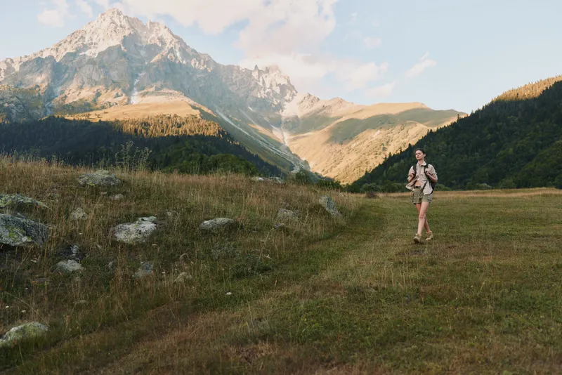 Hiker walking across a meadow with distant mountains, backpack on, bright sunlight, conveying outdoor adventure and calm nature. The moment captures exploration and air.