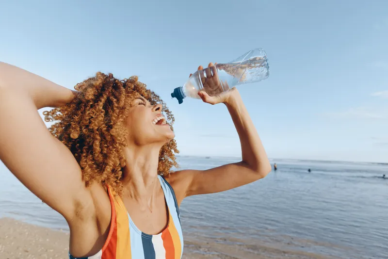 A happy young woman with curly hair drinks water from a bottle on a sunny beach. She wears a colorful striped swimsuit and enjoys the clear blue sky and sea air.
