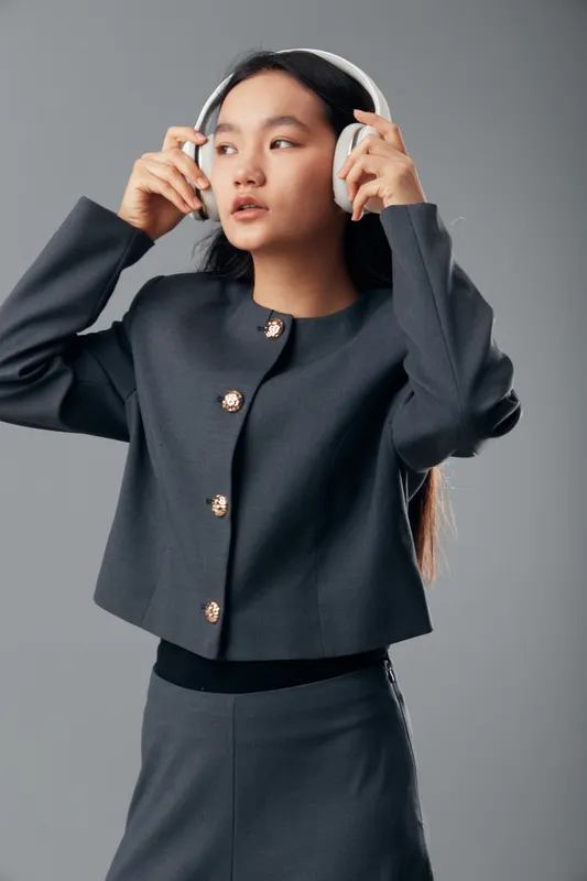Young woman in stylish dark outfit wearing white headphones listening to music, posing on gray background with serious expression and long hair falling on shoulders.