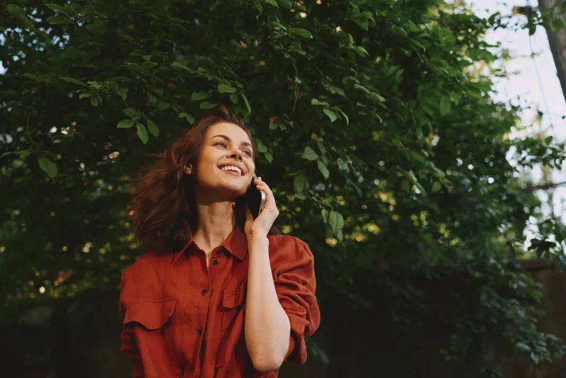 Smiling woman with wavy hair wearing a red shirt standing outdoors, talking on a phone. Natural light, leafy backdrop, casual lifestyle moment focused on connection