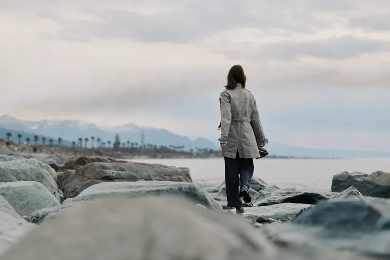 Woman in a gray coat walking alone on rocky beach shore under cloudy sky. Mountains and palm trees visible in distance. Concept of solitude, nature, and contemplation outdoors.