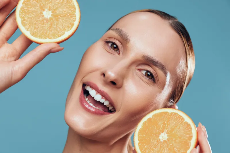 Smiling woman with clear skin holding two halves of fresh orange near her face. Close-up on blue background. Concept of healthy lifestyle, vitamin C, skincare, and wellness.
