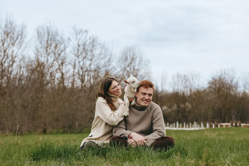 Cheerful couple outdoors, cozy session with a playful goat kid, sunlit grass, warm sweaters, relaxed smiles, candid moment, natural colors, tranquil rural setting, gentle breeze, lifestyle portrait