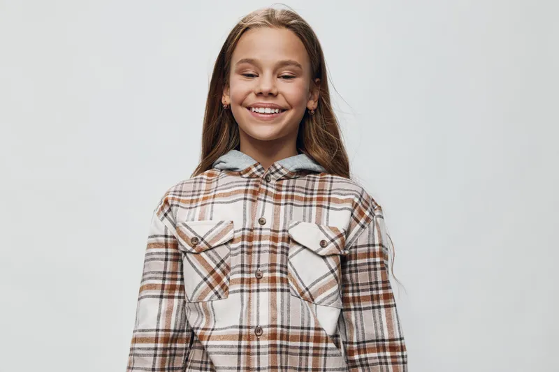 Girl in plaid shirt smiling with closed eyes on plain background. Child with long hair standing relaxed wearing casual checkered jacket with buttons and pockets.