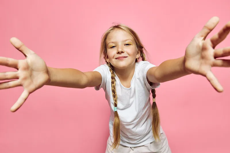 Happy girl with braided hair wearing a white t-shirt reaching forward with open arms on a pink background. Studio portrait with cheerful and playful concept for childhood and joy.