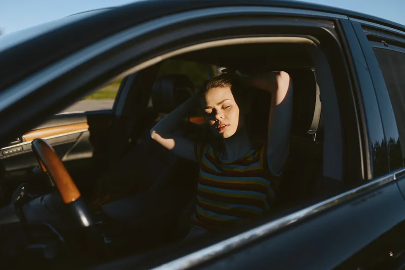 A distressed woman sits in a car with head in hands, overwhelmed by stress. Bright interior, daylight, and retro film color emphasize fatigue and burnout in a candid lifestyle moment.