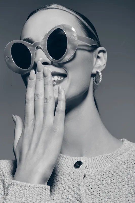 Portrait of a smiling young Caucasian woman with pale skin wearing round sunglasses and hoop earrings covering her mouth with her hand in a stylish knitted sweater.