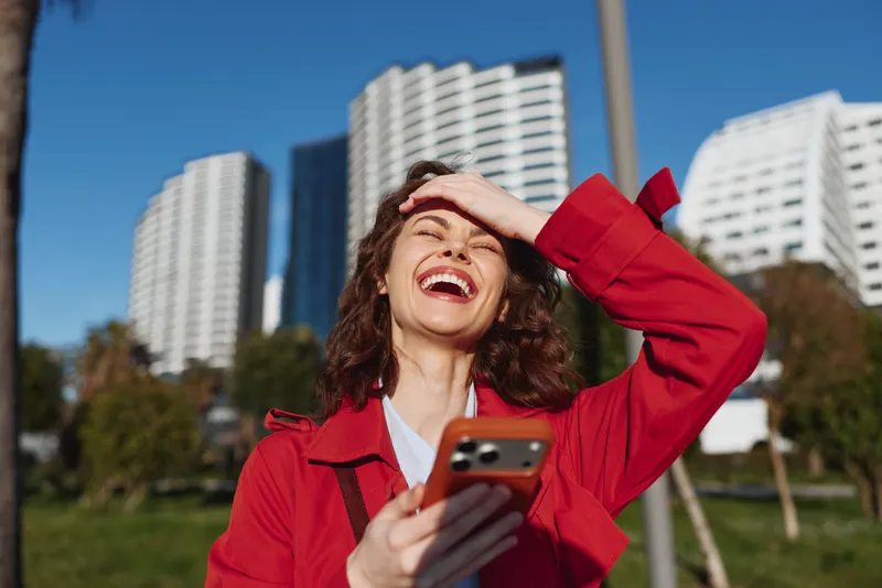 A young woman in a red jacket laughs while shielding her eyes from the sun, holding a smartphone in a bright urban park, conveying spontaneity and carefree mood amid modern architecture.