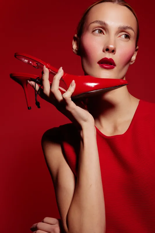 Elegant woman in red dress holds red high heel shoe near face on red background. Fashion concept with bold makeup and stylish accessories for glamour and beauty.