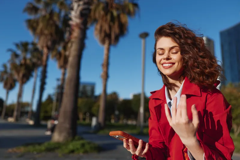A cheerful young woman with curly hair wearing a red jacket holds a smartphone outdoors, smiling with eyes closed in a sunny urban park with palm trees in the background.
