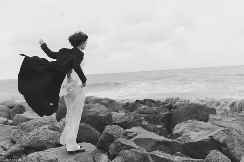 Sea, rocky shore, flowing coat silhouette in black and white, wind-swept motion as a figure stands on stones, dramatic balance and solitude by the coast