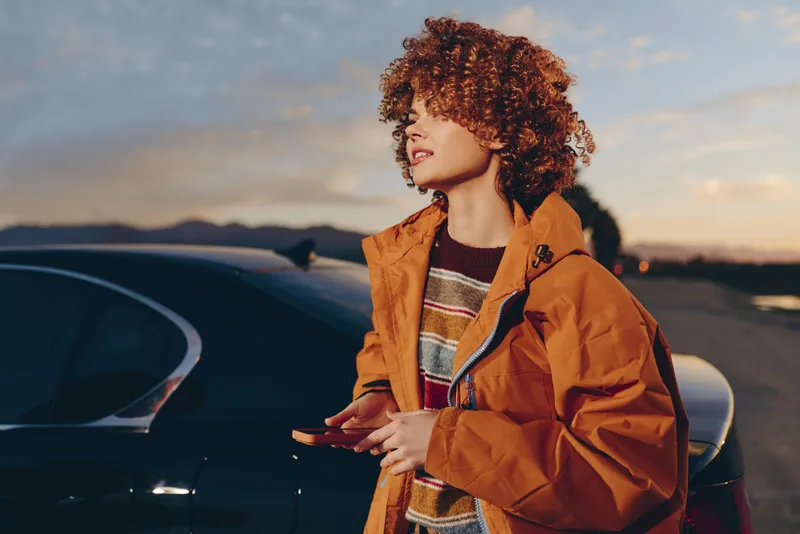 Woman with curly hair stands beside car at sunset, wearing orange jacket, casual lifestyle outdoors, confident profile portrait, wind moving hair, cinematic warm soft light, travel mood