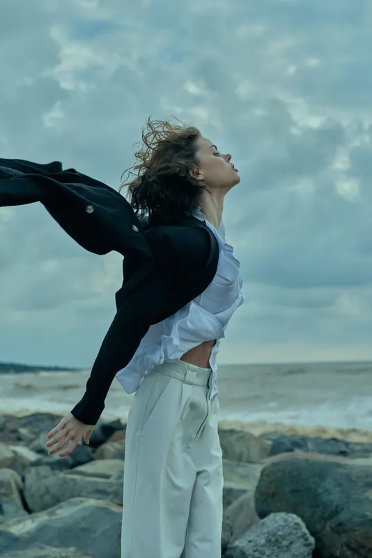 Woman on a rocky beach enjoys wind and motion, clothing billowing as sea and sky form a vast backdrop, an outdoor fashion moment full of freedom