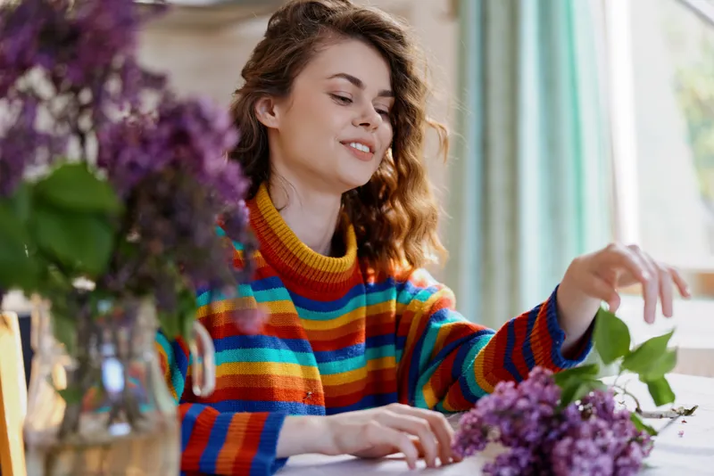 A woman in a colorful striped sweater delicately arranges purple flowers on a bright table, capturing a cozy home moment of hobby, calm, and creative floral craft.