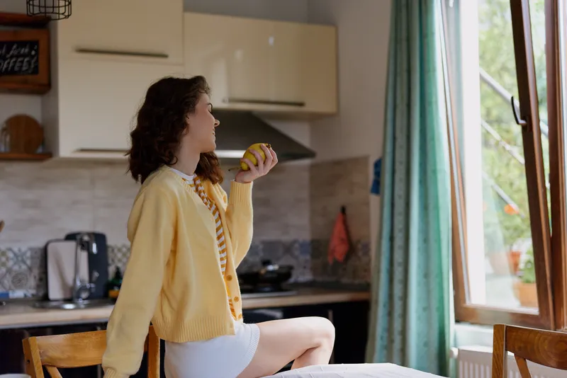A woman in a yellow cardigan sits on a kitchen counter, holding a lemon, bathed in natural window light. A candid home lifestyle moment showcasing casual relaxation and morning warmth.