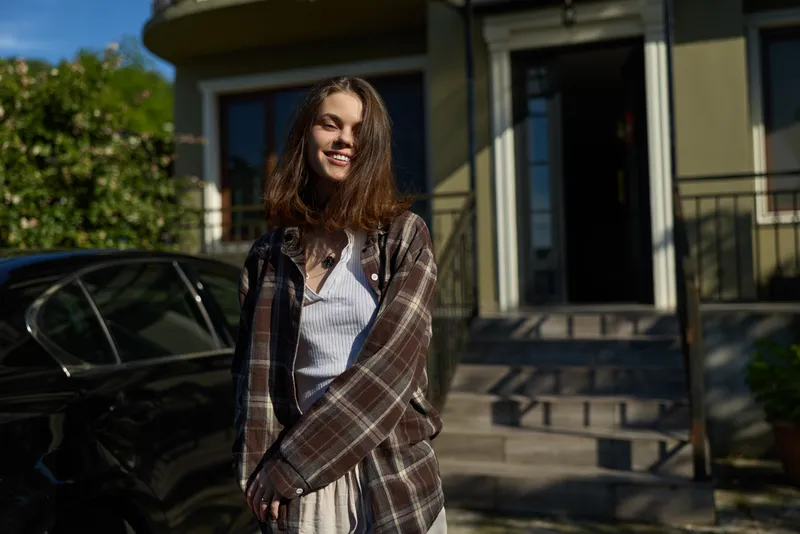 A smiling woman in casual outerwear stands outside a modern house on a sunny day, wrapped in a plaid shawl. A bright lifestyle moment emphasizing relaxed confidence