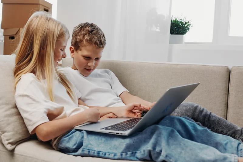 Two children sitting closely on a sofa, focused on a laptop screen. Casual clothing and relaxed posture create a cozy home learning or entertainment concept.
