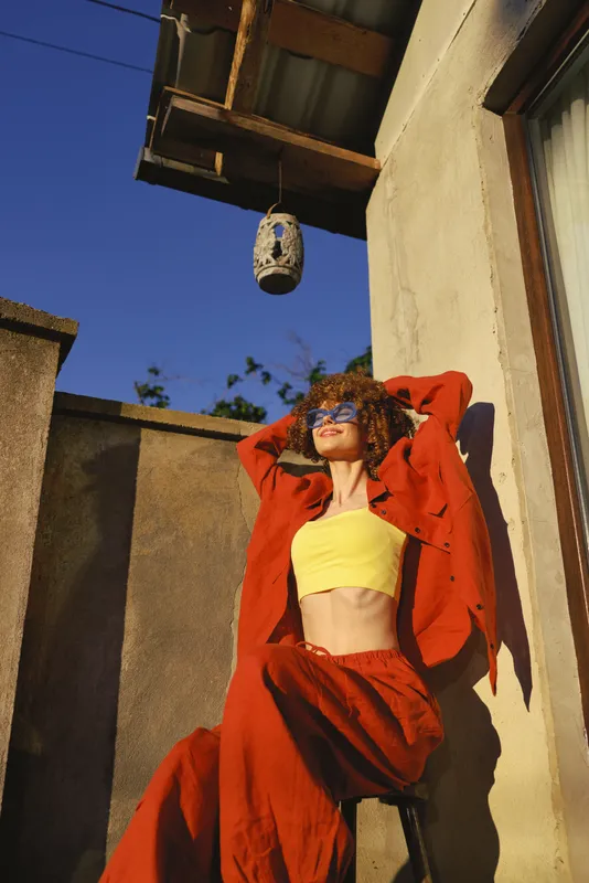 A young woman in a yellow crop top and red jacket lounges on a sunlit rooftop, arms raised and a carefree smile lighting her face
