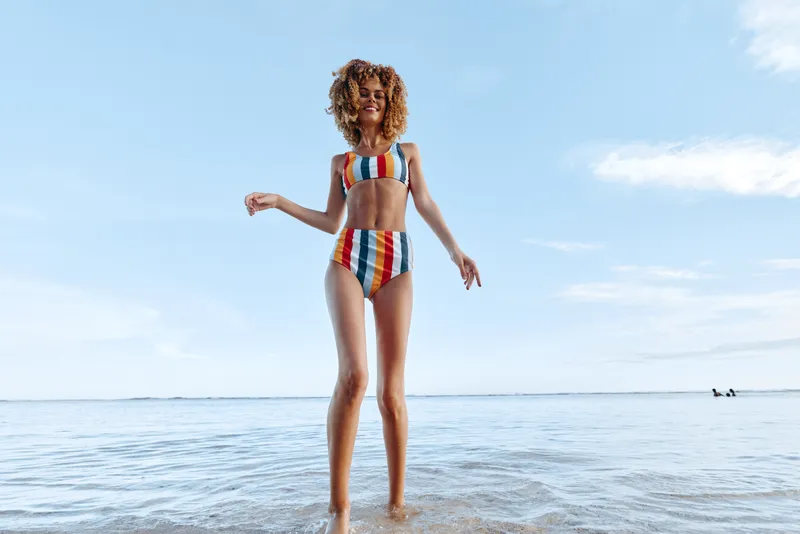 Young woman with curly hair stands on white sand at the edge of the ocean, wearing a rainbow bikini and enjoying a sunlit moment as waves touch her feet, her relaxed pose radiates joy