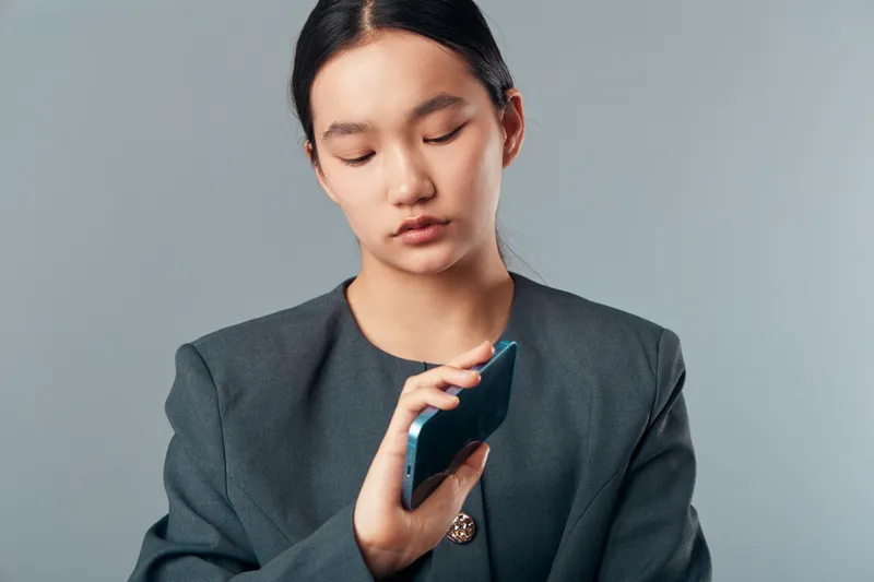 Young woman in professional outfit looking down at a smartphone in hand with neutral gray background. Concentrated female employee using mobile device for texting or reading message.