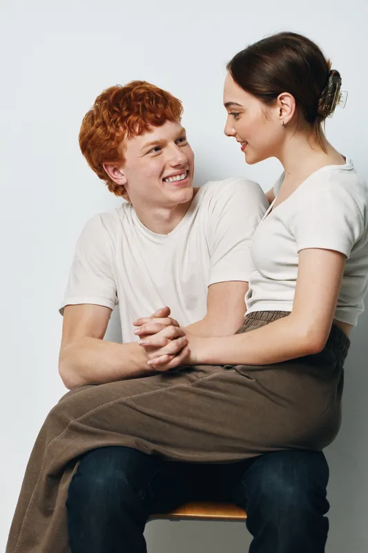 Young couple sitting and smiling at each other in casual clothing indoors. Man with red hair holding woman on lap showing affection and joy in lifestyle scene.