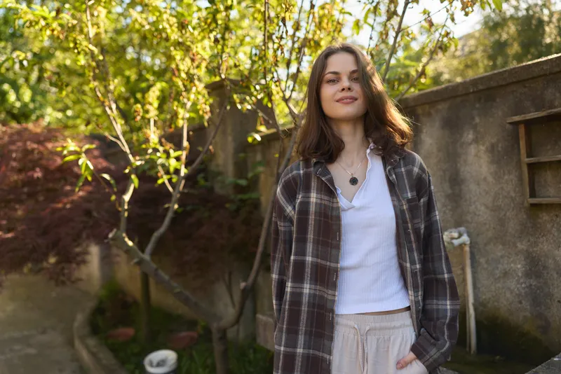 A relaxed woman stands in a sunlit garden wearing a plaid coat over a white top, conveying casual lifestyle and peaceful mood