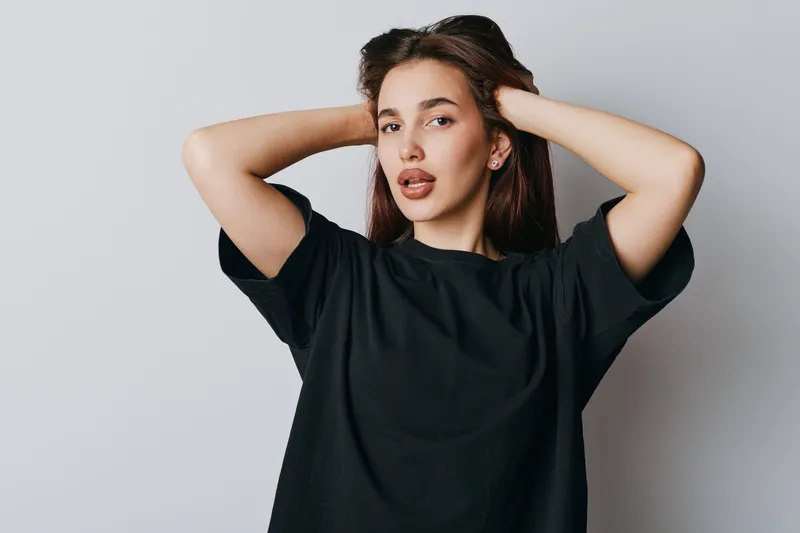 Young woman posing with hands behind head wearing black t shirt against light background. Casual style, confident expression with natural makeup and long dark hair in studio portrait.