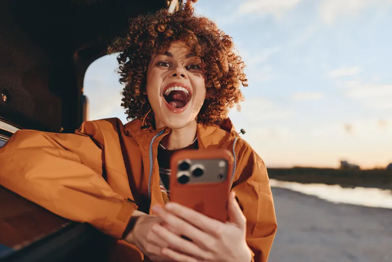 Woman lifestyle selfie smartphone beach sunset curly laughter outdoors, smiling with curly hair in an orange jacket, holding phone toward the camera, near the shoreline, warm evening light