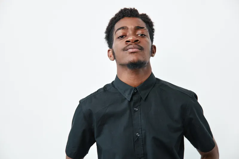 Young black man with dark skin wearing black shirt standing confidently against white background. Portrait of African ethnicity male with short curly hair and serious expression.