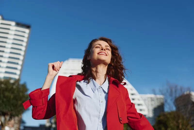 Woman in a red coat stands outdoors, smiling toward the sun with a clear blue sky and city buildings behind, conveying confidence and carefree mood.