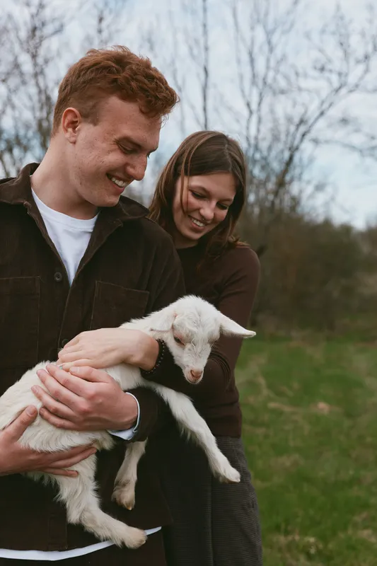 A joyful couple outdoors holds a fluffy lamb, smiling as they cradle the young sheep in a green field, capturing a candid lifestyle moment of gentle animal care and love