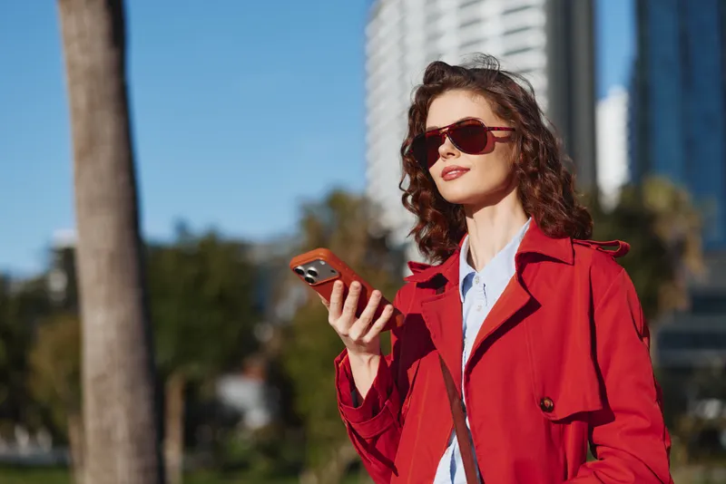 Young woman with curly hair and sunglasses holds a smartphone while walking outdoors in a red coat on a sunny day with urban buildings in the background.