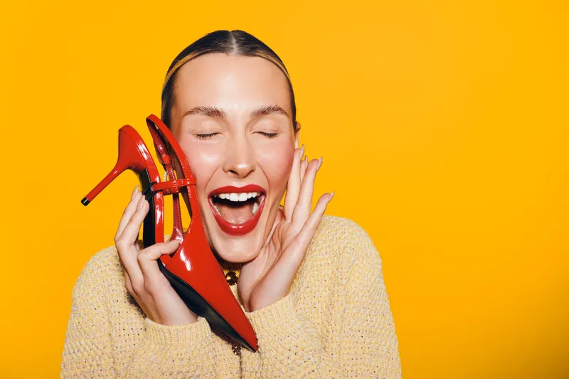 Happy woman with closed eyes holds red high heel shoe near face, smiling with excitement. Bright yellow background and casual sweater create cheerful, vibrant mood and fashion concept.