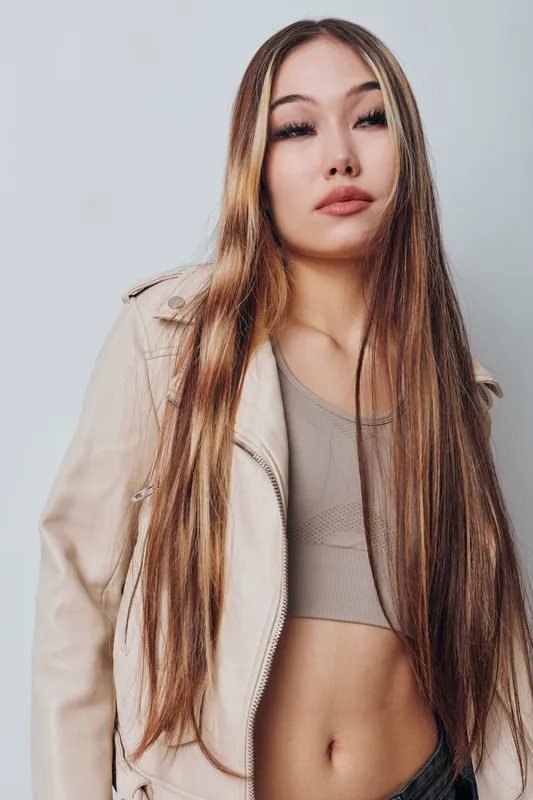 Woman with long straight hair wearing light jacket and crop top posing on gray background for fashion portrait with confident expression and natural makeup.