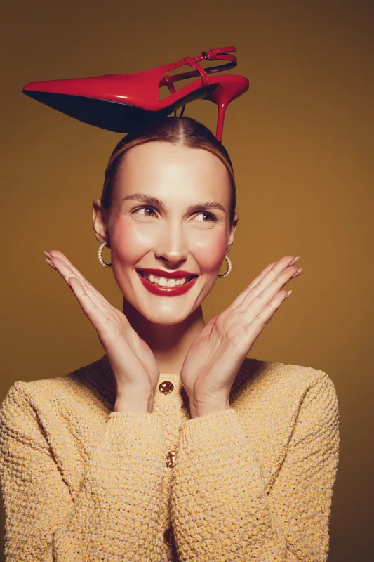 Smiling woman with red high heel shoe balanced on her head, wearing yellow textured sweater and gold hoop earrings. Concept of fashion, fun, creativity, and playful style in studio lighting.