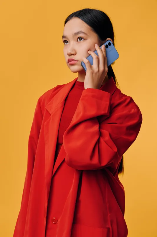 Young woman in red coat talking on smartphone looking away thoughtful on orange background. Portrait, communication, technology, fashion, casual style and modern device in hand.