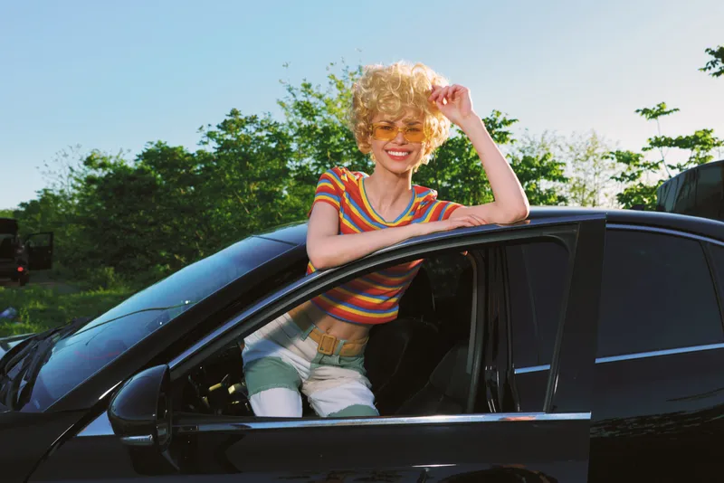 A young woman with curly blonde hair leans out of a sleek black car, smiling under bright sun in a striped casual top, conveying spontaneous road trip joy and carefree summer vibes.